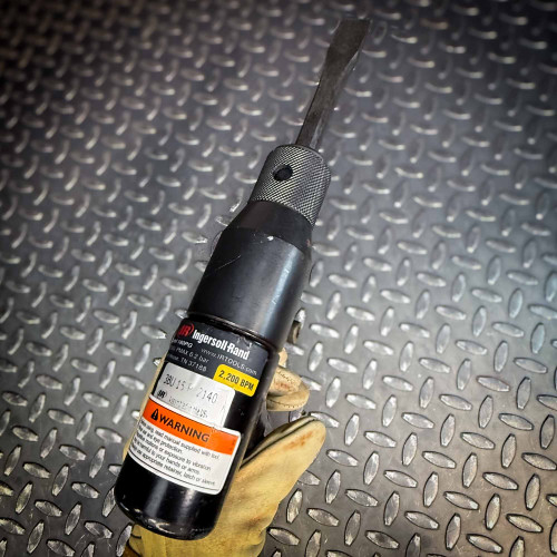 A close-up of the side view of the Ingersoll Rand 180PG Pistol Grip Chisel Scaler on a diamond-plate surface, showcasing the handle and warning labels.