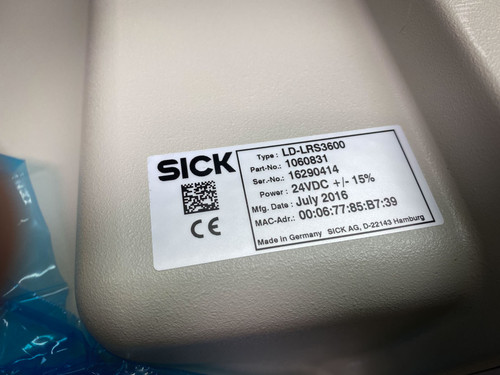This image shows a close-up of a label on a SICK LD-LRS3600 long-range LiDAR sensor. The label includes crucial details such as the type (LD-LRS3600), part number (1060831), serial number (16290414), power requirements (24V DC ±15%), and the manufacturing date (July 2016). Additionally, the label contains a QR code, the CE mark indicating compliance with European standards, and the MAC address of the device. This label confirms the sensor’s specifications and manufacturing origin from SICK AG, Hamburg, Germany, underlining its authenticity and traceability.