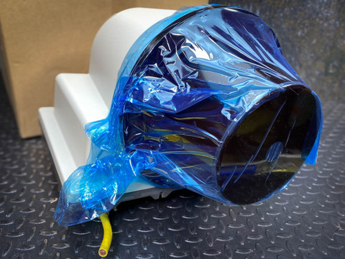 This image presents a close-up view of a SICK LD-LRS3600 long-range LiDAR sensor, a 360° LiDAR scanner. The sensor is shown with its lens end covered in a blue plastic protective wrapping, reflecting a meticulous approach to safeguarding the equipment during handling or shipping. A visible cable, with its yellow and green wires, emerges from the base, hinting at the connectivity requirements of the device. Positioned against a white foam stand and a cardboard box on a textured metal surface, the setup underscores the careful packaging and protection of high-tech equipment in an industrial context.