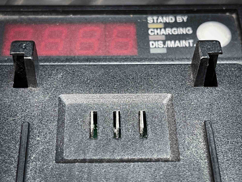This image features a close-up of one of the charging bays in a Kenwood walkie-talkie battery charger. The digital display above the bay shows "8888", suggesting either a standby or diagnostic mode. Below the display, the charger slot has exposed metal connectors for inserting a battery. The labels "Stand By", "Charging", and "Dis./Maint." are visible to the right, indicating the various modes available on the charger. The detailed view emphasizes the technical and functional aspects of the device, set against a gritty, textured background.