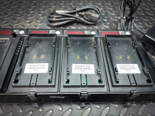This image showcases a close-up view of a multi-bay battery charger designed for Kenwood walkie-talkie batteries, with specific focus on the empty battery slots. Each slot is marked with a model number "AT-2032" and features small LED indicators. A few power cables are also visible in the background, lying on a diamond plate surface, which adds to the rugged and industrial context of the charger.