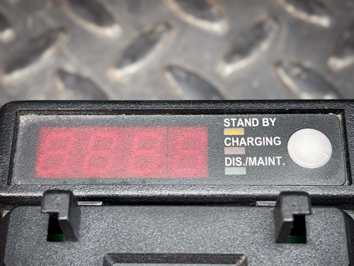The image displays a close-up view of the digital readout and control panel on a battery charger for Kenwood walkie-talkies. The LED display reads "8888" indicating a standby or default screen mode. Next to the display, labels identify the mode buttons: "Stand By", "Charging", and "Dis./Maint." (Discharge/Maintenance), highlighting the charger's multifunctional capabilities. The overall scene has a durable and technical ambiance, emphasized by the rugged background texture of a diamond plate surface.