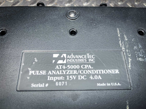 The image offers a close-up of the manufacturer's label on an Advancetec AT4-5000 CPA battery charger. It details the device as a "Pulse Analyzer/Conditioner" with technical specifications of "Input: 15V DC 4.0A". The serial number "6071" and "Made in U.S.A." are also prominently displayed. The label is set against the backdrop of a worn black casing with visible scratches and paint wear, indicating heavy usage. The metallic screws and surface textures enhance the industrial look of the equipment.