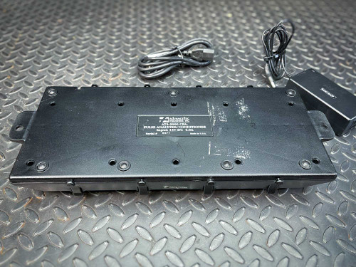 The image features the underside of an Advancetec battery charger for Kenwood walkie-talkies, model AT4-5000. This view highlights the product label that specifies it as a "Pulse Analyzer/Conditioner" with an input specification of 15V DC and 4.0A. Wear marks and scratches on the black casing indicate frequent use. Two power cords are positioned beside the charger on a textured, diamond plate surface, suggesting readiness for connection and deployment in a technical or industrial environment.