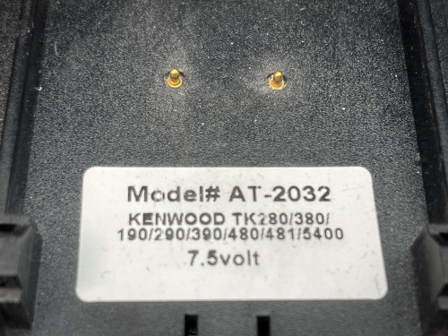 This image shows a close-up view of the label on a battery charger bay designed for Kenwood walkie-talkies. The label displays "Model# AT-2032" and lists compatible Kenwood models such as TK280, TK380, TK190, TK290, TK390, TK480, TK481, TK5400, all operating at 7.5 volts. The surrounding area of the label is textured and speckled with dust, highlighting its rugged use environment. Two small LED indicators above the label suggest the operational status of the charger.