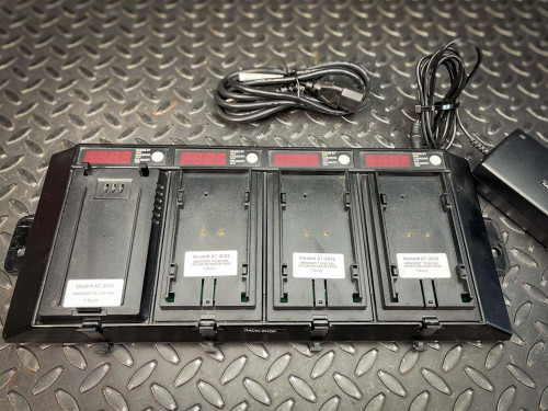 The image shows a multi-slot battery charger specifically designed for Kenwood walkie-talkie batteries, model AT-2032. There are several battery slots, each with a digital display showing the charging status. The textured metal surface beneath adds a durable and industrial feel to the setting. Cables and a power adapter are visible on the right side of the image, indicating the setup is ready for use.