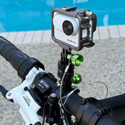 Close-up view of a GoPro Mount action camera attached to a bicycle handlebar, secured with a safety tether. The camera is an Insta360 GO 3, held by a metallic frame with green and black adjustment knobs. The background shows a swimming pool. GoPro Mount action camera safety tether from Obtainium Science and Surplus.