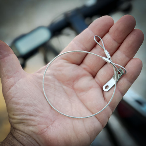 A hand holding a safety tether loop, designed for securing a GoPro Mount action camera. The background is blurred, showing parts of a bicycle. GoPro Mount action camera safety tether from Obtainium Science and Surplus.