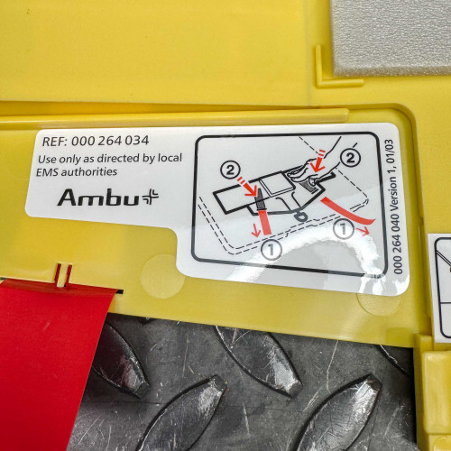 The image features another instructional panel on the Ambu Head Wedge cervical immobilization device, highlighting the application steps for proper use. The diagram, marked with steps 1 and 2 and red arrows, illustrates how to correctly place and secure the device around a patient's head. The text on the panel reads "Use only as directed by local EMS authorities" and includes the Ambu logo and reference number, emphasizing the device's medical and emergency use context.