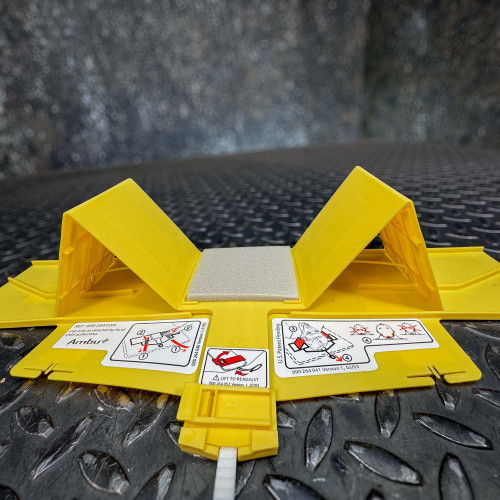 This image captures a wider perspective of the Ambu Head Wedge cervical immobilization device set against a reflective, textured metal surface. The device is displayed in a partially unfolded state with yellow panels and integral white labels showing detailed usage instructions. Diagrams on the labels depict steps for adjusting and securing the head straps, marked with red arrows for clarity. This setup highlights the practical, instructional aspects of the device, ensuring correct application during emergency medical scenarios.