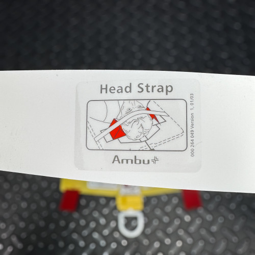 The image displays a close-up of an instructional panel on the Ambu Head Wedge cervical immobilization device, focusing on the "Head Strap" usage. The diagram shows a schematic of a head with a red strap securing it, demonstrating proper head stabilization technique. The panel includes the Ambu logo and a reference number, emphasizing the medical context and functionality of the device.