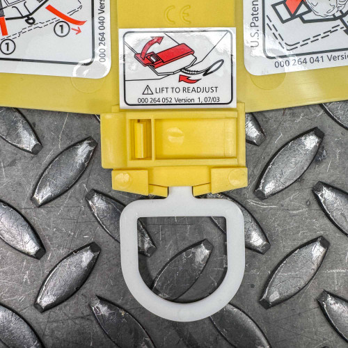 The image showcases a detailed close-up of a specific component on the Ambu Head Wedge cervical immobilization device. It features a diagram on a white label illustrating the step "Lift to Readjust," which shows how to adjust a clip on the device for proper fit. The background is a yellow section of the device with the same diagram, and it is positioned above a textured metal surface. The photo captures the practical details intended to guide the correct handling of the device.