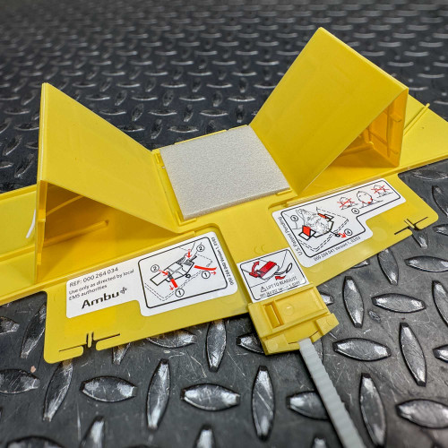 The image displays the Ambu Head Wedge cervical immobilization device in a partially folded position on a textured metal surface. Visible are several yellow panels, one of which has a grey padding attached, and instructional diagrams detailing steps for correct application. The diagrams include icons indicating the adjustment of straps and proper positioning, labeled with reference numbers and warnings to use only as directed by local EMS authorities. This view emphasizes the device's functional design and its readiness for use in medical emergencies.
