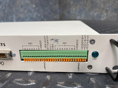 A detailed view of the National Instruments BNC-2090 terminal block's green and orange connector section, placed on a textured metal surface. Available from Obtainium Science & Industry Surplus.