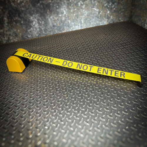 A yellow retractable barrier labeled 'CAUTION - DO NOT ENTER' from Crowd Control Warehouse, neatly extended on a dark textured metal floor, from Obtainium Science and Surplus.