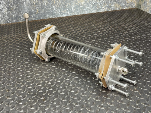 Full view of the Obtainium Science and Surplus custom-made vacuum/pressure chamber with a cylindrical glass body, internal coil, and metal end plates on a textured metal surface. Multiple bolts secure the ends, showing sturdy construction.