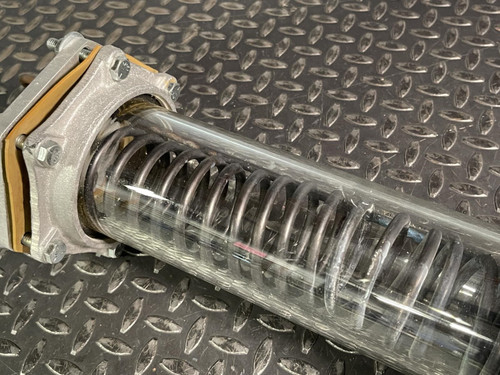 Angled close-up of the Obtainium Science and Surplus vacuum/pressure chamber, showing the transparent body and internal coil. The metal end plate and bolts are visible, emphasizing the durability of the custom-built component.