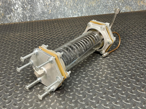 Full image of the Obtainium Science and Surplus vacuum/pressure chamber, featuring the glass tube, internal heat-exchange coil, and metal plates at both ends with secured bolts, resting on a textured black metal surface.