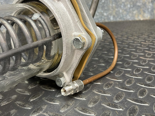 Detail shot of the Obtainium Science and Surplus vacuum/pressure chamber’s connection points, showing the metal end plate, layered gaskets, and attached copper tubing, resting on a dark metal surface with a textured pattern.