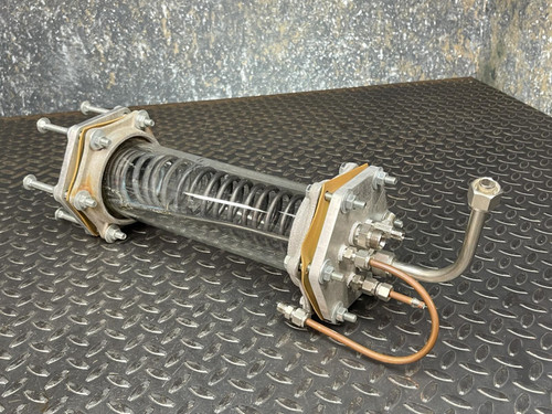 Custom-made vacuum/pressure chamber with an internal heat-exchange coil from Obtainium Science and Surplus. The chamber is displayed on a textured, dark metal surface, featuring a cylindrical glass body, metal end plates, and attached copper tubing.