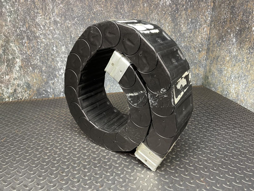 A coiled Nylatube Energy Chain Wireway, displayed against a textured metal surface. The wireway is black with a slightly reflective finish, showcasing its flexible and durable construction.