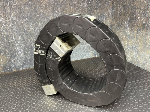 A coiled Nylatube Energy Chain Wireway, available from Obtainium Science & Industry Surplus, displayed against a textured metal surface. The wireway is black with a slightly reflective finish, demonstrating its flexibility and durability.