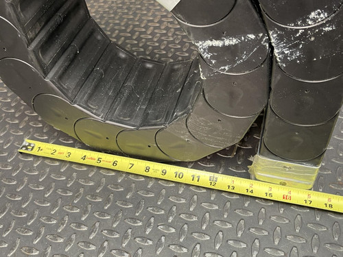 Close-up of a coiled Nylatube Energy Chain Wireway with a yellow tape measure placed below it for scale, showing measurements in inches. The wireway, available from Obtainium Science & Industry Surplus, is displayed on a textured metal surface, highlighting its flexibility and dimensions.