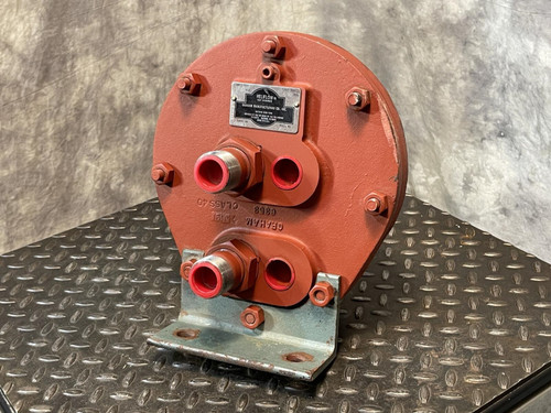 Graham heat exchanger from Obtainium Science and Surplus, shown upright on a textured black metal surface. The red metal end plate has multiple bolt fittings and two central ports, indicating it’s designed for industrial use.