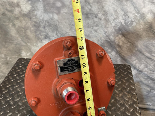 Front view of the Graham heat exchanger from Obtainium Science and Surplus, with a yellow measuring tape placed vertically across the red end plate to indicate height. The component has bolts and ports, resting on a textured metal surface.