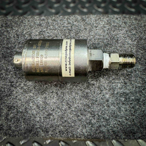 Image of an OmegaDyne pressure transducer model PX01D1-5KGV, showing its details including the serial number 159329 and a pressure range of 0-5000 PSI. The label also includes the contact information "www.omegadyne.com" and "info@omegadyne.com." The device is used for measuring pressure and is placed on a textured surface with a metal background.