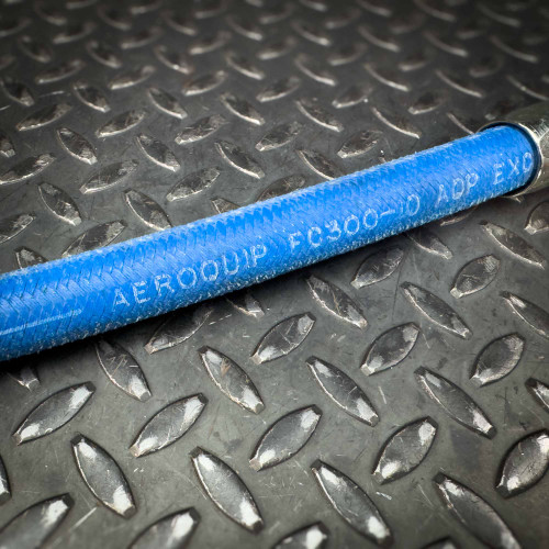 A detailed view of the blue Aeroquip FC300 hose from Obtainium Science and Surplus, focusing on the printed text along the hose's surface, with the textured metal background in contrast.