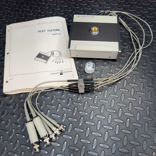 A 1970s vintage Hewlett Packard 16021A Test Fixture for the HP 4271A LCR Meter, shown with several cables and connectors, and an operating manual on a textured surface.