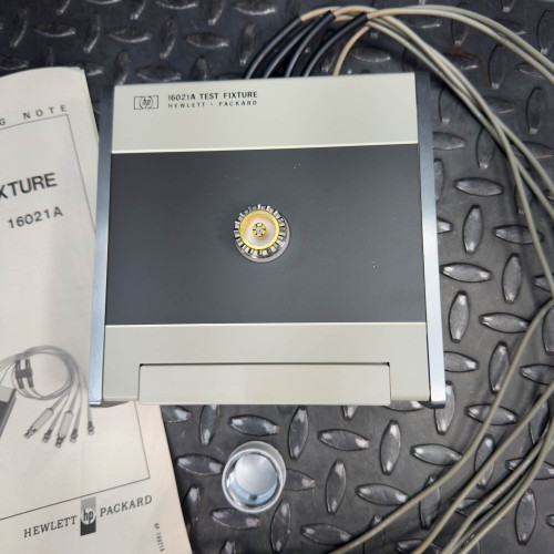 Top view of the Hewlett Packard 16021A Test Fixture showing the main device with cables extending from it, and an adjacent operating manual.