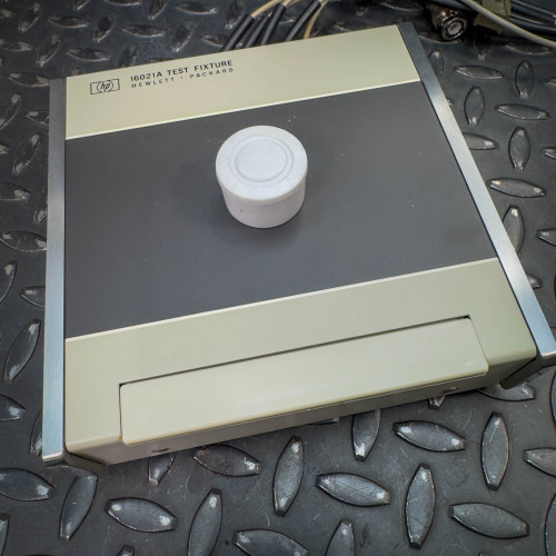 The Hewlett Packard 16021A Test Fixture with a white cap covering the central connector, placed on a textured surface.