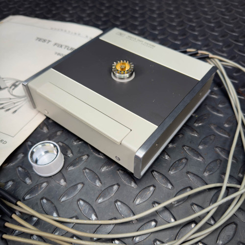 Close-up of a Hewlett Packard 16021A Test Fixture showing the central component with connected cables and a partially visible manual.