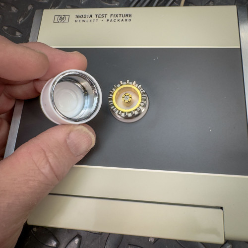 A hand holding a part of the Hewlett Packard 16021A Test Fixture, showing a close-up of the  cap that protects the central  connector of the fixture.