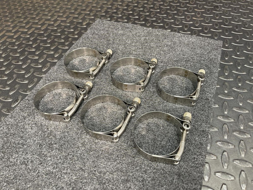 Six Clampco 2.5" stainless T-bolt clamps arranged neatly on a gray textured surface. These clamps, sourced from Obtainium Science and Surplus, have a smooth metallic finish, and their robust construction is ideal for heavy-duty applications.