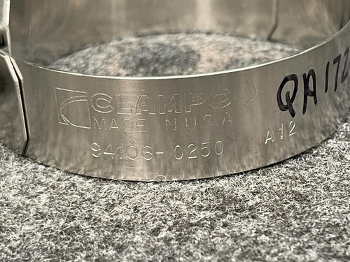 A focused shot of the side of a Clampco 2.5" stainless T-bolt clamp, showing the embossed product details along the band. Visible text reads part numbers and specifications, with "QA 172" written in marker, indicating a quality inspection from Obtainium Science and Surplus.