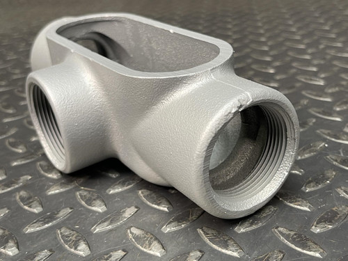 Close-up of a T conduit body from Obtainium Science and Surplus, focusing on its threaded ports and oval opening on a metal surface.