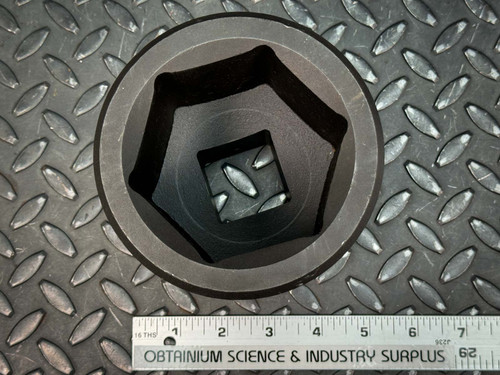 Top view of a black metal socket with a hexagonal interior and a square drive hole, placed on a textured metal surface with a ruler showing a length of approximately 3.5 inches. This socket is from Obtainium Science & Industry Surplus.