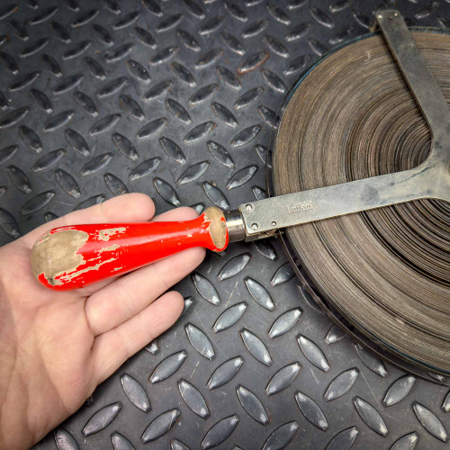 This image features a close-up of a person's hand holding the handle of a Lufkin 254TC steel gauging tape measure, which extends from a large metal reel containing the coiled 300-foot tape. The handle is painted red with visible wear and chipped paint, emphasizing frequent use. The backdrop is a diamond plate metal surface, enhancing the industrial aesthetic of the scene.