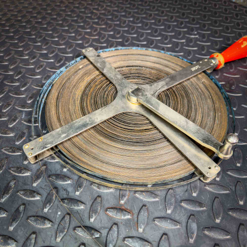 This image depicts a Lufkin 254TC steel gauging tape measure coiled on a round metal reel, with a large, cross-shaped winding mechanism positioned centrally. The reel sits on a textured diamond plate metal surface, emphasizing the tool's rugged, industrial environment. The edge of the metal tape is partially visible, and the wind-up handle has a red grip with visible signs of use, suggesting frequent operation in a professional setting.