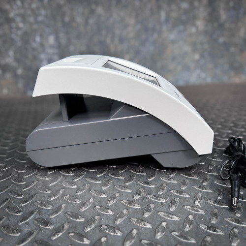 The image provides a side view of the Siemens CLINITEK Status+ Analyzer, showing its sleek, modern design. The analyzer is featured with a two-tone color scheme, consisting of a light gray top and a darker gray base, which gives it a professional and contemporary look. The ergonomic curve of the device not only adds aesthetic appeal but also suggests functionality and ease of use in a clinical setting. The power cable in the image suggests readiness for connection and use, and the sturdy base ensures stable operation on various surfaces, like the textured metal shown here.
