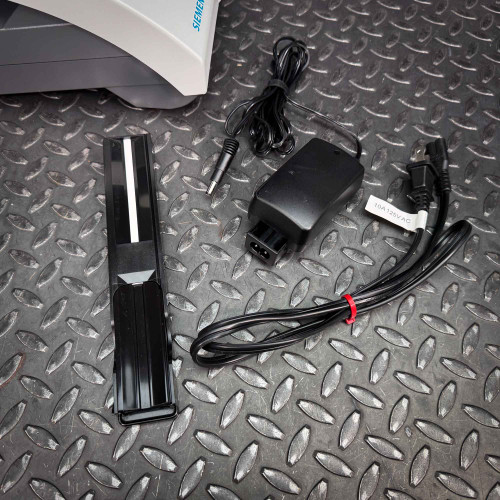 The image displays accessories associated with the Siemens CLINITEK Status+ Analyzer, laid out on a textured metal surface. Included are the power supply adapter, a black power cord, and a removable black sample cassette tray. The tray appears sleek and is designed for easy handling and insertion into the analyzer. This setup is typical for clinical environments, where each component is essential for the operation of the diagnostic device, ensuring it functions efficiently for urine analysis tests.