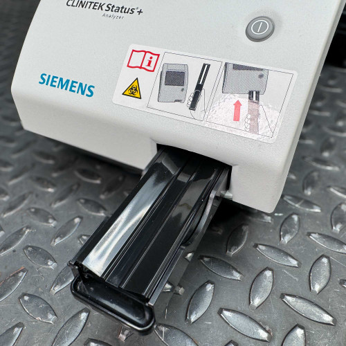 The image zooms in on a specific feature of the Siemens CLINITEK Status+ Analyzer, showcasing a slide-out, removable cassette tray where test strips or samples are likely inserted for analysis. The drawer is partially extended, revealing its glossy black surface. Above the drawer, you can see the analyzer’s instructional diagram, which provides visual guidance on how to insert a test strip correctly. It is highlighted by a warning symbol and the operational button. This view highlights the practical design and operational focus of the analyzer, set against a textured metal background indicative of a clinical setting.
