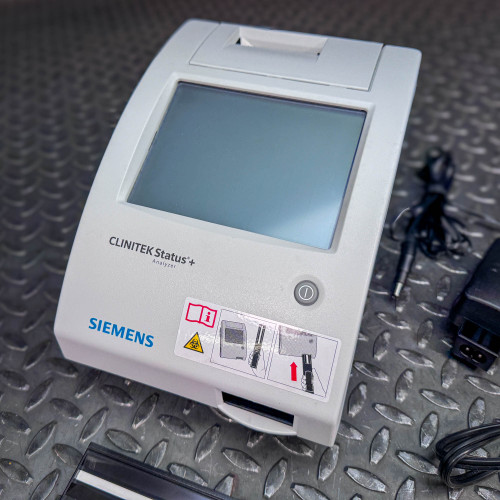 The image provides a closer view of the Siemens CLINITEK Status+ Analyzer, focused on the front panel with its large screen and operational buttons. The analyzer's design is compact and user-friendly, featuring clear branding and model identification. A diagram with warning symbols and operational instructions is also visible on the front, guiding the user on proper usage. The background features a textured metal surface, suggesting a robust, clinical or laboratory environment suitable for such medical equipment.