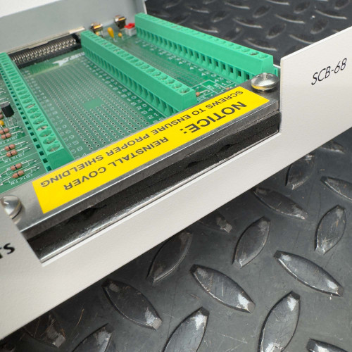 The image shows an angled view of the open National Instruments SCB-68 terminal block connector. The interior reveals green circuit boards and connection points, with a prominent yellow label stating "NOTICE: Remove Shield Before Using" and "REINSTALL COVER BEFORE USING." The device is placed on a textured metal surface.
