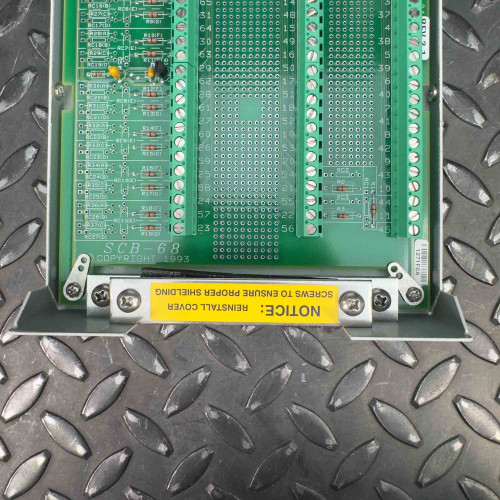 The image provides a close-up view of the bottom part of the interior of a National Instruments SCB-68 terminal block connector. It focuses on the green circuit board with its numerous connection points and the yellow label that reads, "NOTICE: Remove Shield Before Using." The background is a textured metal surface.