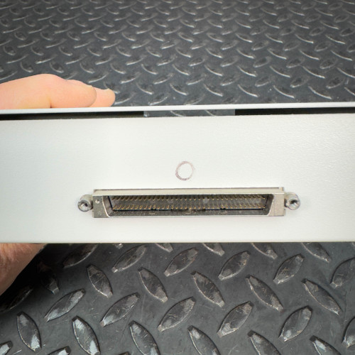 The image shows a close-up of the back side of a National Instruments SCB-68 terminal block connector, highlighting the connector port. The rectangular device is placed on a textured metal surface, and a hand is partially visible holding the device. The connector port is centrally located, with mounting screws on either side.