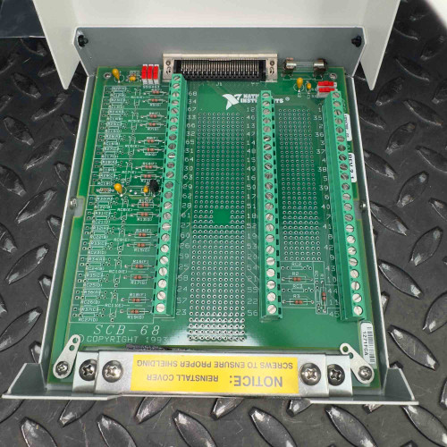Internal view of a National Instruments SCB-68 I/O terminal block connector with the cover removed, revealing the green circuit board and various electronic components, placed on a black textured metal surface. The device is available from Obtainium Science & Industry Surplus.